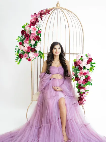 A beautiful and regal portrait. The mom-to-be is seated in a golden birdcage chair adorned with flowers, wearing a flowing lavender gown.