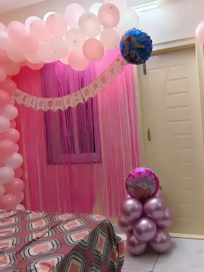 A full view of a bedroom decorated for a baby girl's arrival. The pink shimmer curtain backdrop, balloon arch, and "Welcome Baby" banner create a perfect photo opportunity and a festive atmosphere.