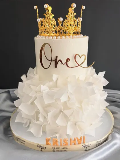 A full view of the elegant all-white ruffled dress cake, crowned with a golden 'One' topper for a truly special first birthday.