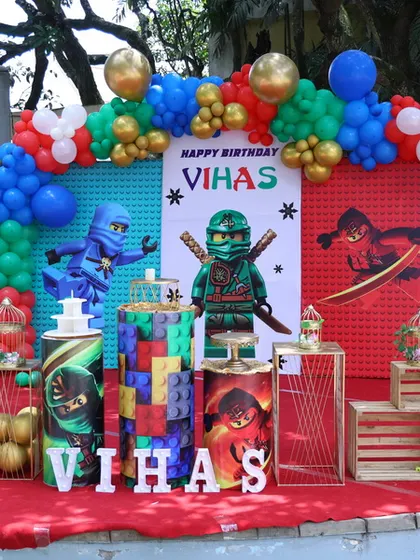 A complete birthday party setup for a Ninjago theme, featuring a custom backdrop, character displays, themed plinths for cake and desserts, and a vibrant balloon arch.