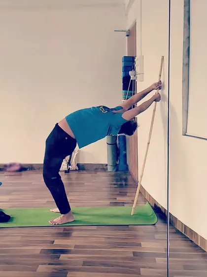 A student uses the wall and a stick to work on a standing backbend, a great way to build confidence and flexibility safely.