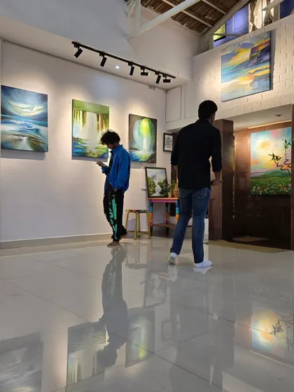 Two visitors explore the gallery during Gurpreet Kaur's solo show, surrounded by her vibrant and ethereal landscape paintings.