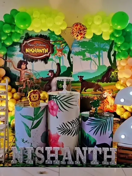 A Jungle Book themed birthday party with a vibrant backdrop showing Mowgli and Baloo. The setup is decorated with tropical leaf-printed pedestals and a large marquee number '2'.