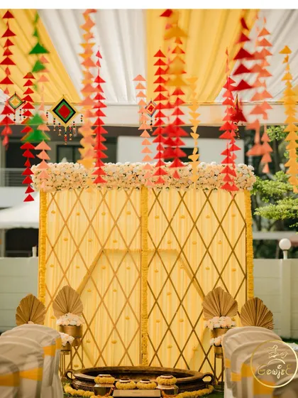 A beautiful Haldi setup with a yellow lattice backdrop, floral arrangements, and hanging paper crafts.