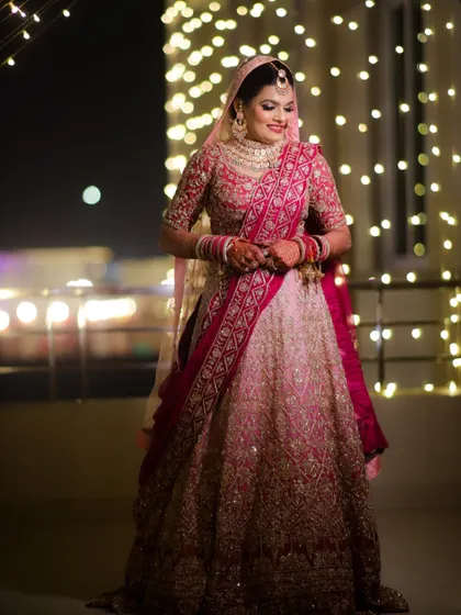 A full-length shot of a bride in her pink lehenga, smiling against a backdrop of lights. Her makeup is glowing and radiant, perfect for an evening ceremony.