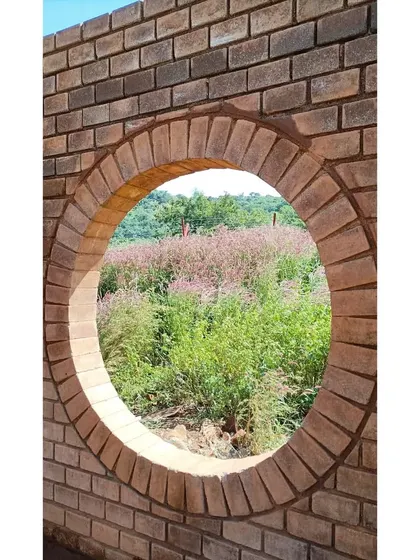 A circular opening in a newly built brick wall at the Gamaya site frames the vibrant wildflowers beyond. We believe in creating architecture that celebrates and connects with the beauty of its immediate environment.