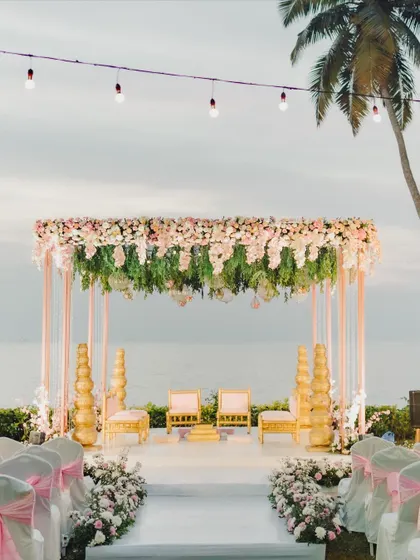 A breathtaking beachside mandap in Goa, with a canopy fatores of pink and white flowers, set against the ocean.
