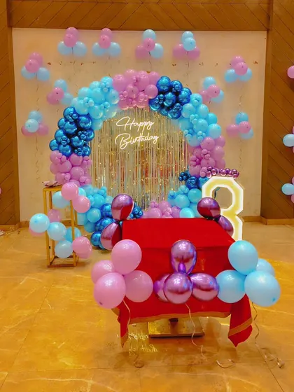 A stylish and modern birthday setup featuring a circular balloon arch with blue, pink, and chrome purple balloons. The shimmering silver backdrop and 'Happy Birthday' neon sign create a perfect spot for photos.