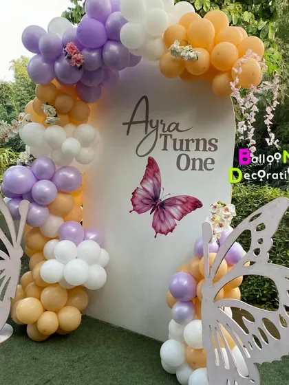 Ayra's butterfly-themed first birthday in an outdoor setting. The decor features large butterfly wing props and a beautiful balloon arch in purple, orange, and white.