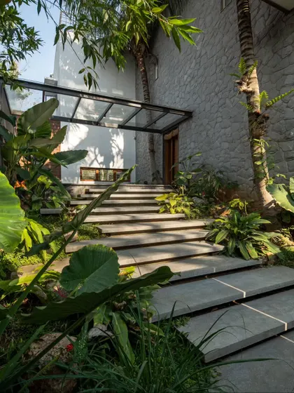 A view of the entrance pathway, where floating concrete steps are surrounded by lush tropical foliage, leading up to the main door under a glass canopy.