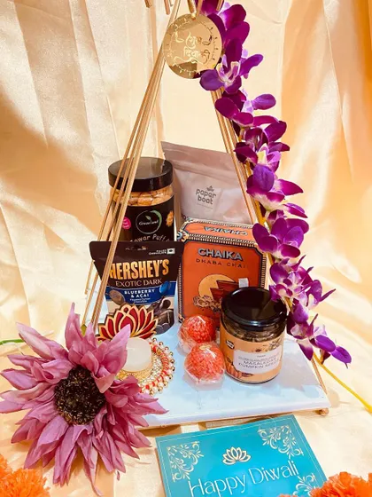 A modern and artistic take on Diwali gifting. This hamper features a unique triangular stand adorned with orchids, holding a curated selection of teas, chocolates, and festive sweets.
