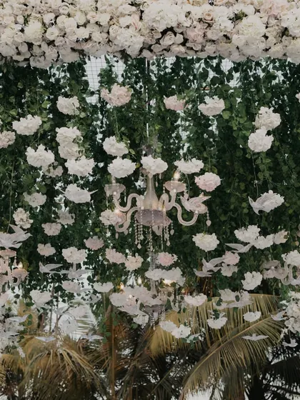 An ethereal, all-white wedding decor theme. The ceiling is adorned with hanging white flowers, paper birds, and elegant crystal chandeliers, creating a dreamy, floating garden effect.