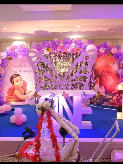 A first birthday party for a baby girl with a fairy and butterfly theme. The stage includes a large butterfly prop, "ONE" marquee letters, and cutouts of fairies and princesses.