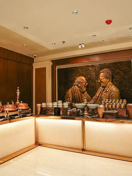 An indoor buffet setup with elegant wood-paneled walls and a striking piece of art in the background. The illuminated counter and copper serving dishes create a warm and luxurious ambiance.