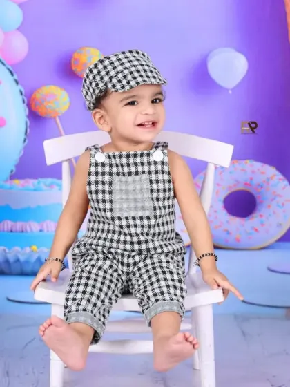 A baby boy in a checkered outfit sits on a white chair in the middle of a pastel sweet shop. His happy expression shows he's having a wonderful time.