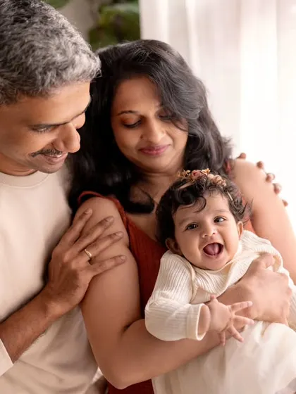 A family of three enjoying a moment by the window. The baby's happy laugh and the parents' loving smiles are beautifully illuminated by natural light.