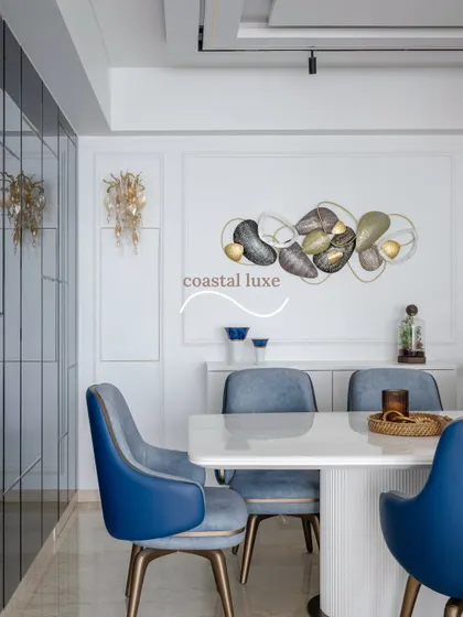 An example of coastal luxe design in a dining room, where a white marble-top table is paired with sapphire blue chairs and ocean-inspired art. White paneled walls and a mirrored accent wall enhance the light, airy feel.