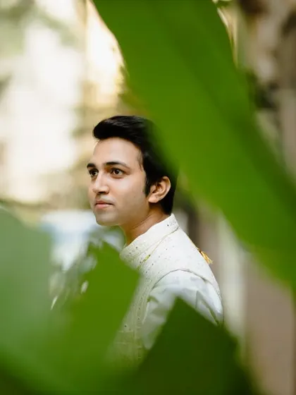 A handsome portrait of the groom, captured through a frame of green leaves. His thoughtful expression and traditional attire are highlighted in this creative and well-composed shot.