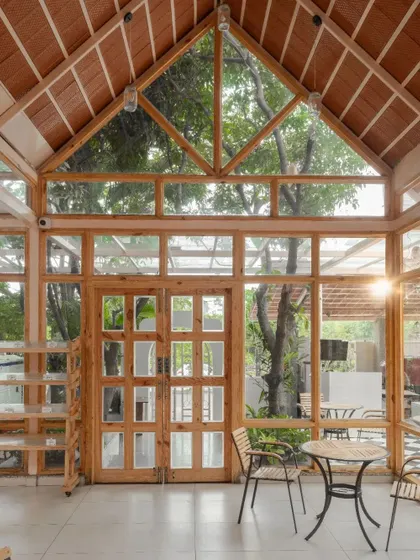 The main dining hall features a high, pitched roof and glass walls, creating a light-filled, greenhouse-like atmosphere. The wooden structure and large windows frame views of the surrounding trees.