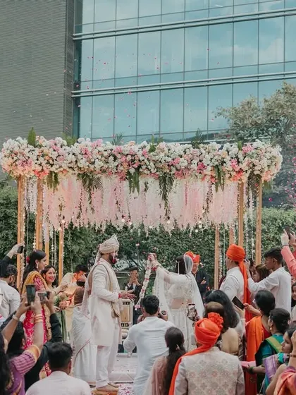 A wide shot of the varmala ceremony taking place at an outdoor mandap, surrounded by guests, showcasing a beautiful day wedding.