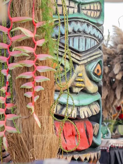 A close-up of a colorful Tiki statue, adorned with hanging pink flowers and surrounded by natural fibers.