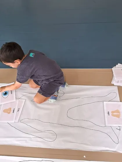 A boy places cards of body parts onto his outline, learning to identify and locate different parts of his body.