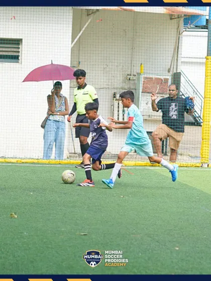 A player shields the ball from a defender under the watchful eye of the referee and parents.