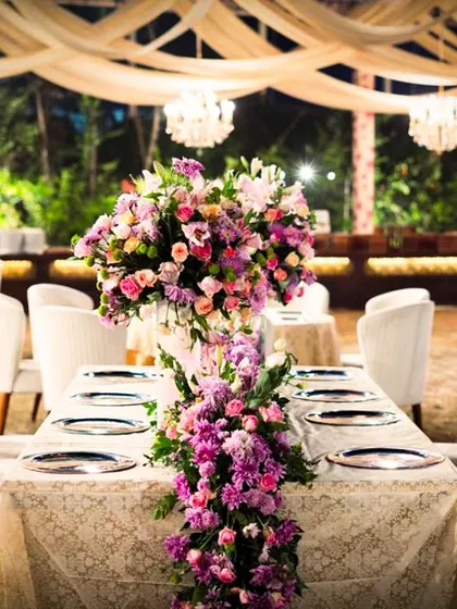 A stunning cascading floral arrangement in shades of purple and pink serves as the centerpiece for this long dining table. The arrangement adds height, drama, and a touch of romance to the reception setting.