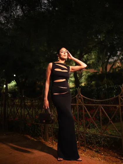 A full-body shot with a pensive, high-fashion pose. The ambient light of the park at night creates a moody and atmospheric backdrop for the sleek black dress.