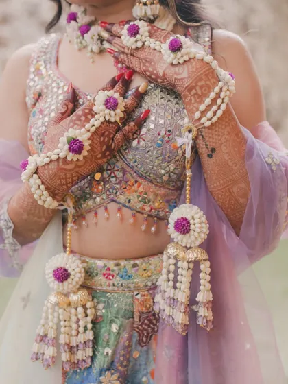 A close-up of the bride's hands, showcasing our intricate haath phool with purple flowers and pearl details, set against her mirror-work blouse.