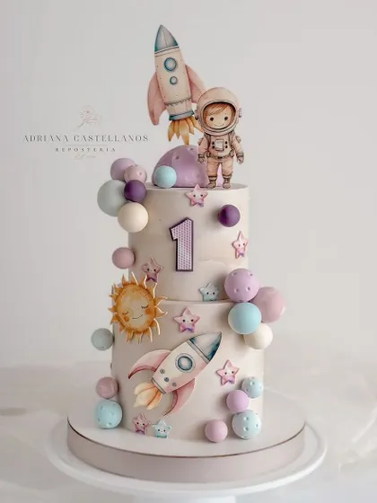 An out-of-this-world space-themed cake for a first birthday. This two-tiered cake features rockets, planets, and a cute little astronaut.