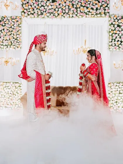 A Varmala ceremony on a stage that seems to float on clouds, thanks to the use of dry ice, with a floral and chandelier backdrop.