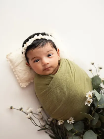 A close-up of a beautiful newborn girl, swaddled in olive green and framed by delicate white wildflowers. This highlights her calm and curious expression.