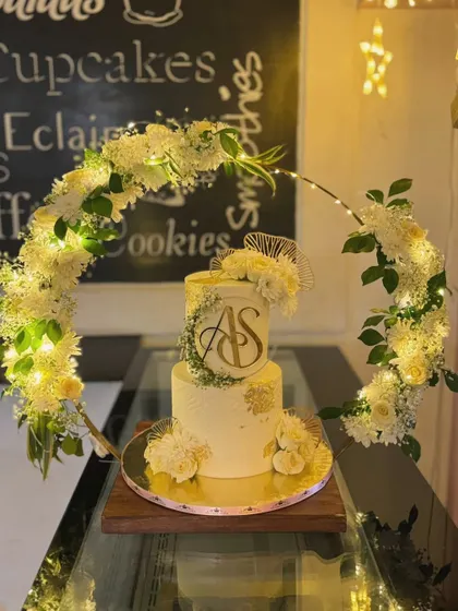 This two-tiered custom wedding cake features a stunning illuminated floral hoop, creating a breathtaking centerpiece for an evening celebration. The design incorporates a gold acrylic monogram, delicate white flowers, and soft lighting for a truly magical effect.