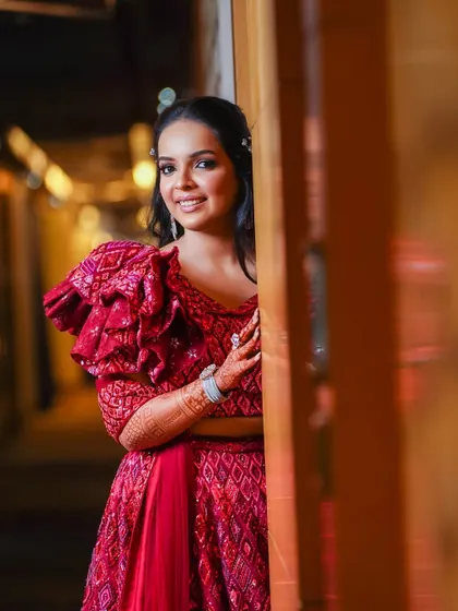 A beautiful portrait of the bride peeking out from a doorway. Her modern red lehenga with ruffled sleeves and her happy smile create a chic and joyful image.