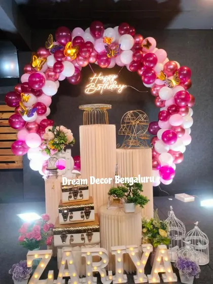 A vibrant birthday setup with a circular balloon arch in shades of magenta and pink. The marquee letters, birdcage props, and plinths create a full and festive look.