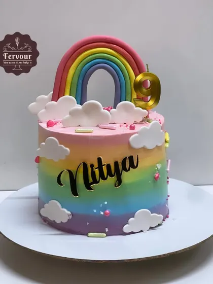 A vibrant rainbow cake with a smooth gradient of colors. The design is topped with a fondant rainbow and clouds, and personalized with a gold acrylic name topper.