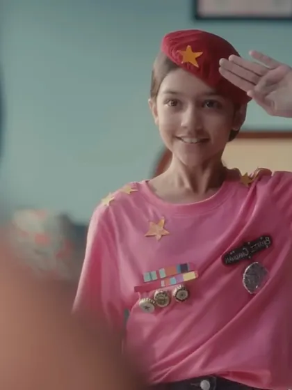A close-up of the young character saluting her reflection. The combination of the red beret and the imaginative, self-made uniform was designed to show her early determination and the start of her journey.
