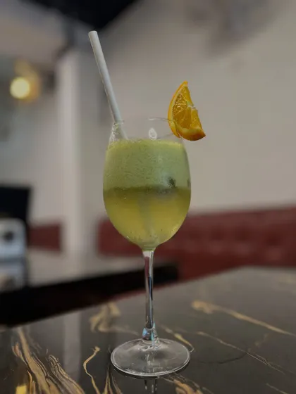 A refreshing green mocktail, likely a kiwi or green apple flavor. The simple composition against the restaurant's interior focuses on the drink's cool color and elegant presentation in a wine glass.