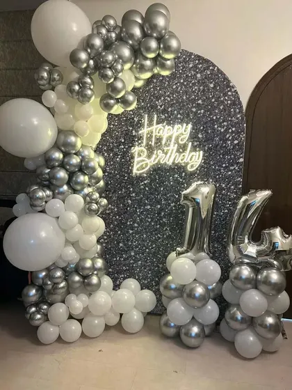 A 16th birthday decoration with a silver glitter backdrop. A white and silver chrome balloon garland and matching foil number balloons complete the chic look.