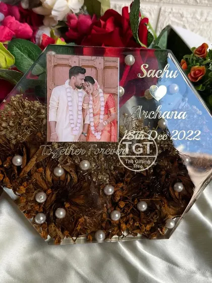 The hexagon shape provides a modern frame for the couple's photo and preserved flowers. The names and wedding date are inscribed in elegant silver text.