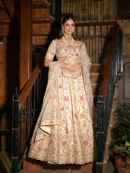 A full-length view of the bride on the stairs. The elegant ivory lehenga is complemented by makeup that features a smokey eye and a natural, glowing complexion.