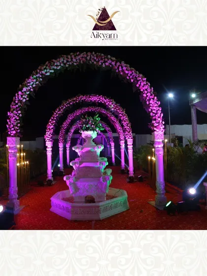 A grand reception entrance with a series of floral arches leading to a beautiful water fountain. The purple uplighting and red carpet create a royal and opulent welcome.