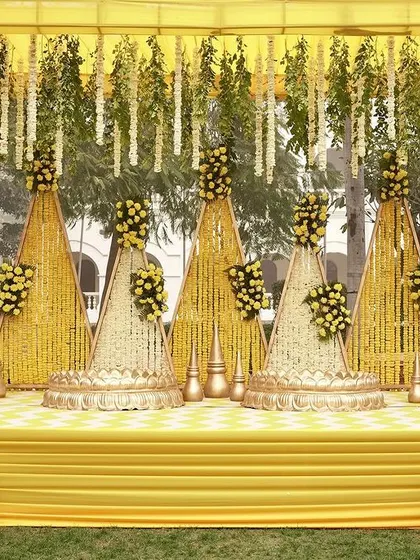 A traditional Haldi stage design with a modern twist. The backdrop features triangular panels covered in marigolds, with hanging floral strings and elegant gold finials.