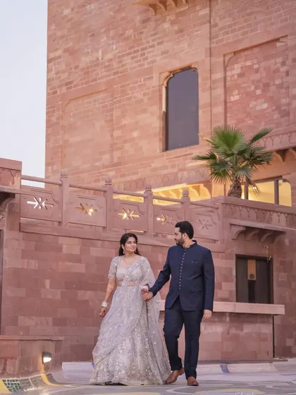 A classic walking shot against the beautiful red sandstone architecture of their Jodhpur hotel, capturing the couple in their elegant traditional attire.