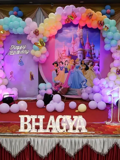 A beautiful pastel princess theme for a first birthday. The stage is decorated with a rainbow balloon arch over a castle backdrop, with character stands of various Disney princesses creating a royal celebration.