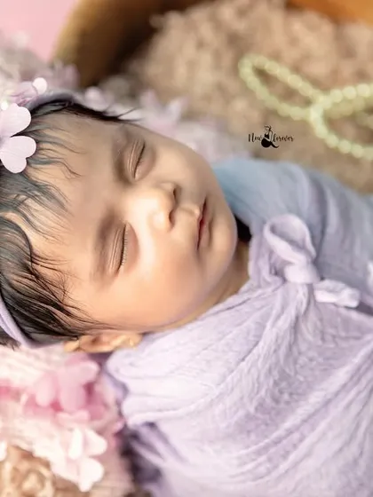 A close-up of a sleeping newborn's face, showcasing her delicate features and peaceful expression. The soft lavender wrap and floral headband complete this angelic look.