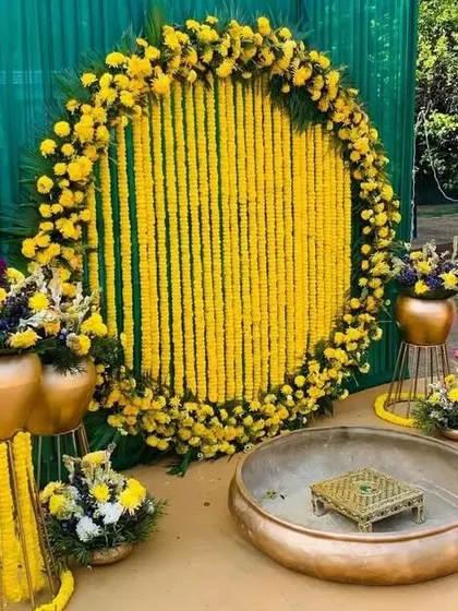 An outdoor Haldi setup with a circular marigold garland backdrop against a green screen. The arrangement includes a traditional brass 'urli' and matching floral arrangements on stands.