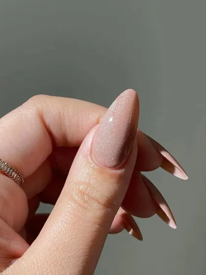 A close-up detail of the shimmering nude polish on a long stiletto nail, showing the fine, reflective glitter within the gel.