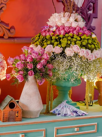 A close-up of a floral installation, showcasing the beautiful mix of pink roses and other blooms. The decor includes charming details like a miniature birdhouse and decorative fans.
